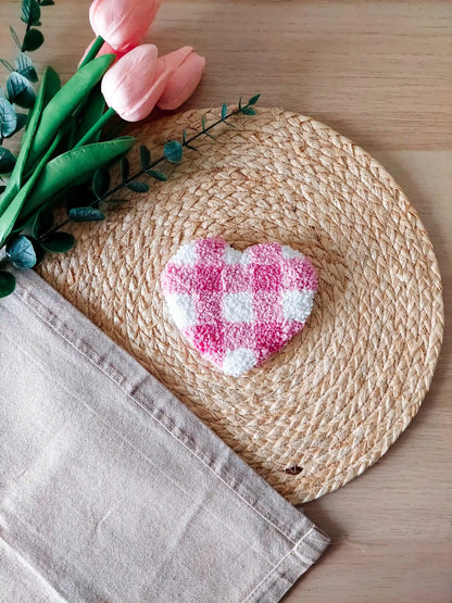 Handmade punch needle heart-shaped mug rug with a pink and white checkered pattern, photographed on a woven mat with decorative flowers in the back. 