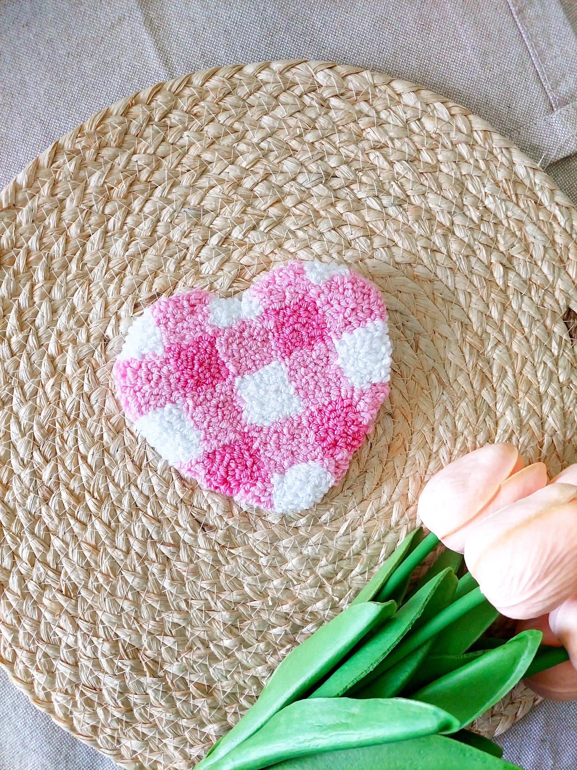Handmade punch needle heart-shaped mug rug with a pink and white checkered pattern, photographed on a woven mat with decorative flowers in the back. 
