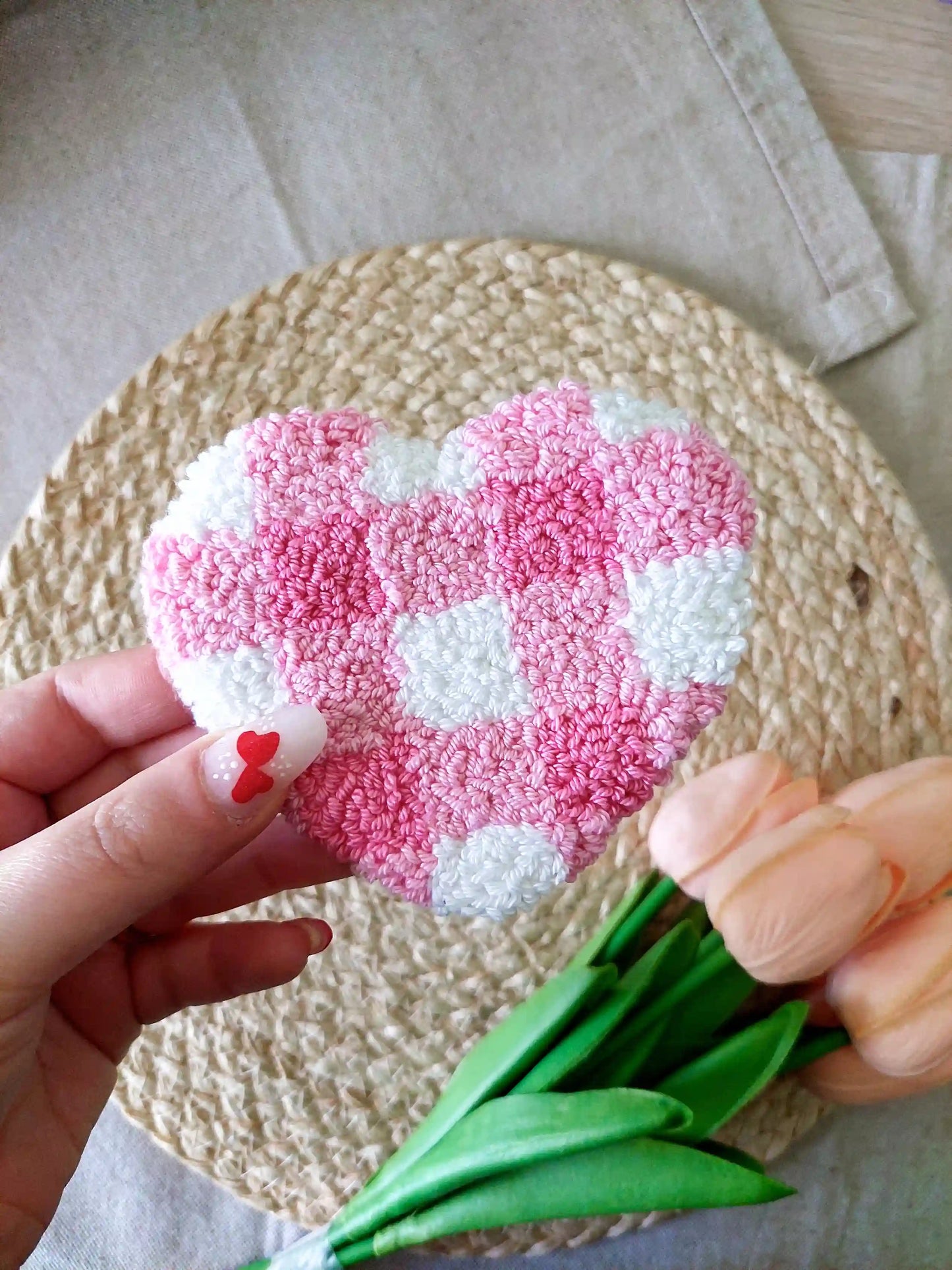 Handmade punch needle heart-shaped mug rug with a pink and white checkered pattern, photographed on a woven mat with decorative flowers in the back. 