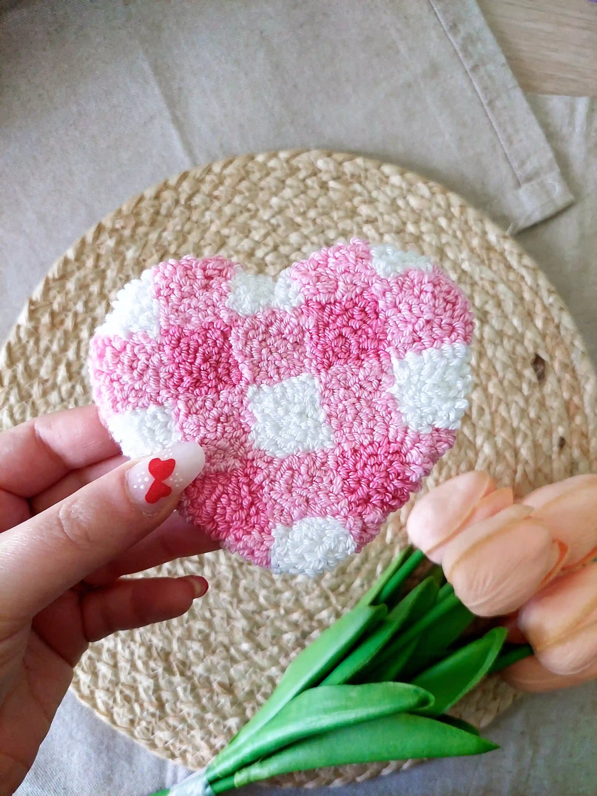Handmade punch needle heart-shaped mug rug with a pink and white checkered pattern, photographed on a woven mat with decorative flowers in the back. 