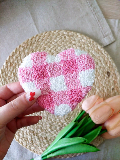 Handmade punch needle heart-shaped mug rug with a pink and white checkered pattern, photographed on a woven mat with decorative flowers in the back. 