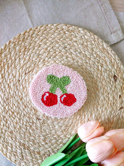 Handmade round punch needle coaster featuring two red cherries with green leaves on a soft pink background, photographed on a woven mat with decorative pink flowers in the back.
