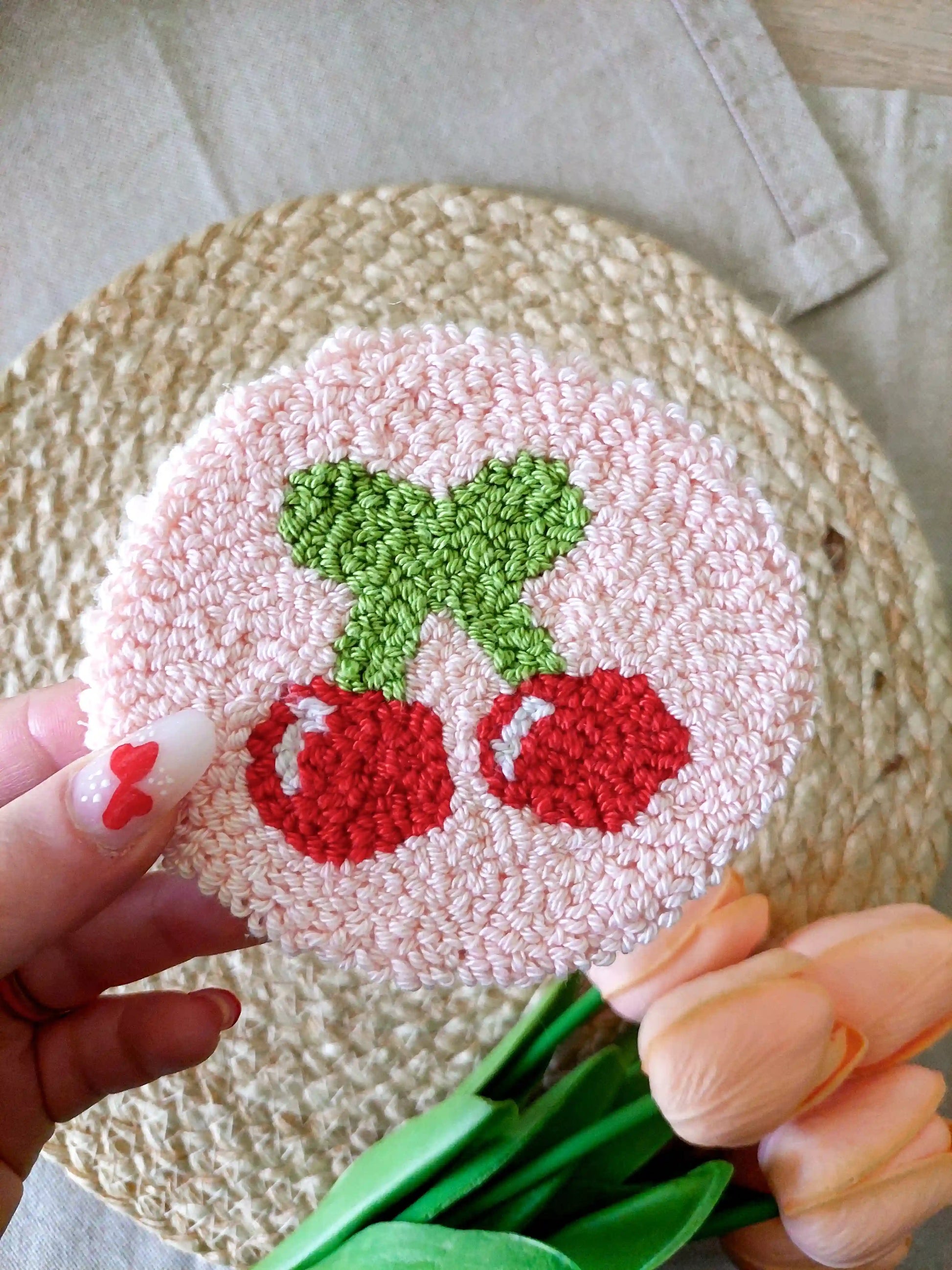 Handmade round punch needle coaster featuring two red cherries with green leaves on a soft pink background, photographed on a woven mat with decorative pink flowers in the back.