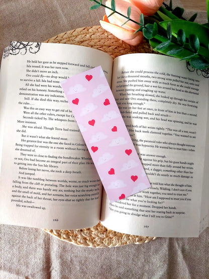 Bookmark featuring a pattern with  smiling clouds and red hearts on a pink background, resting on an open book.