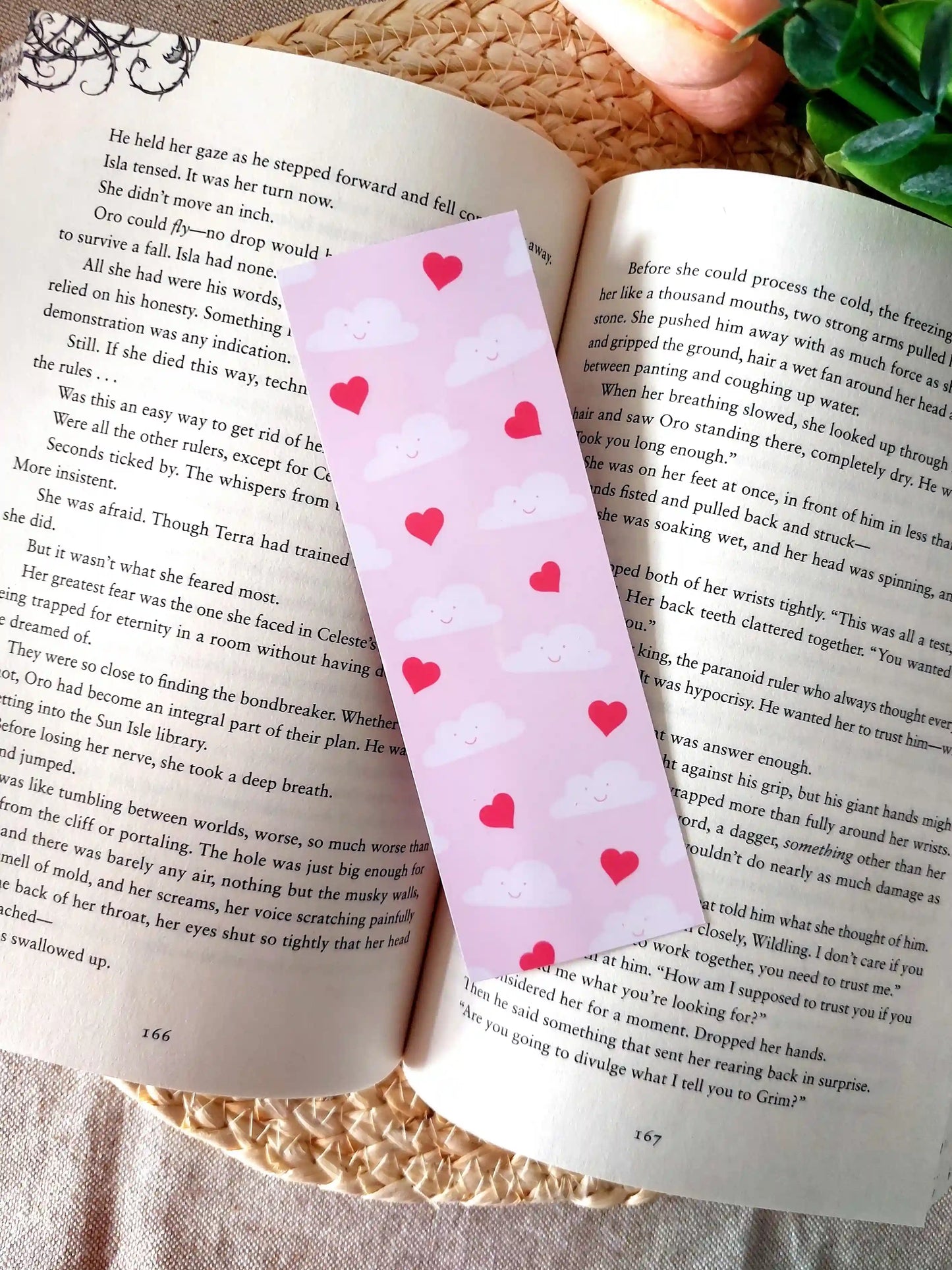 Bookmark featuring a pattern with  smiling clouds and red hearts on a pink background, resting on an open book.