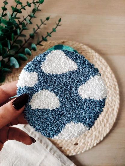 Round blue punch needle coaster featuring white clouds, photographed on a woven mat with decorative flowers in the back.