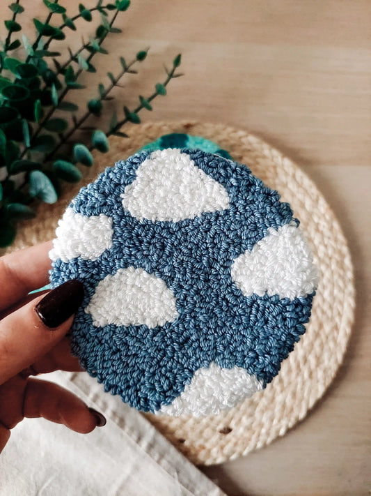 Round blue punch needle coaster featuring white clouds, photographed on a woven mat with decorative flowers in the back.