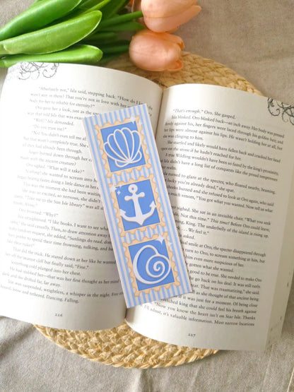 An open book featuring a bookmark with blue beachy stamp-like illustrations (a seashell, anchor and a shell) on a striped blue and white background.