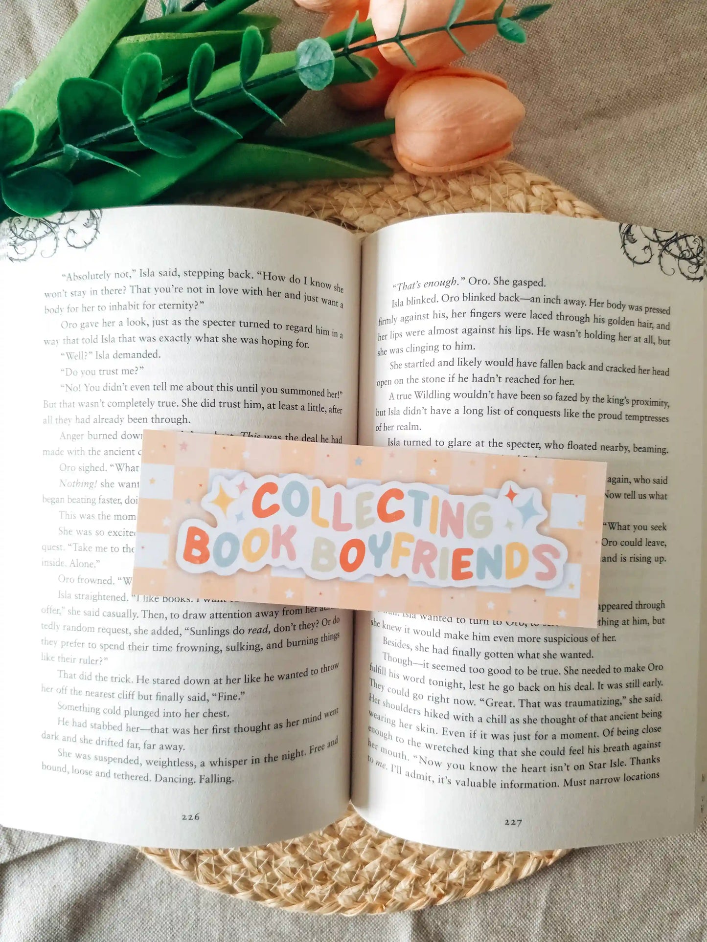 A orange bookmark reading 'COLLECTING BOOK LOVERS' in pastel lettering, laid across an open book. The scene is styled with green leaves and peach flowers on a woven mat over a light fabric background.
