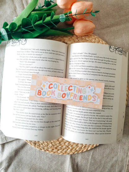 A orange bookmark reading 'COLLECTING BOOK LOVERS' in pastel lettering, laid across an open book. The scene is styled with green leaves and peach flowers on a woven mat over a light fabric background.