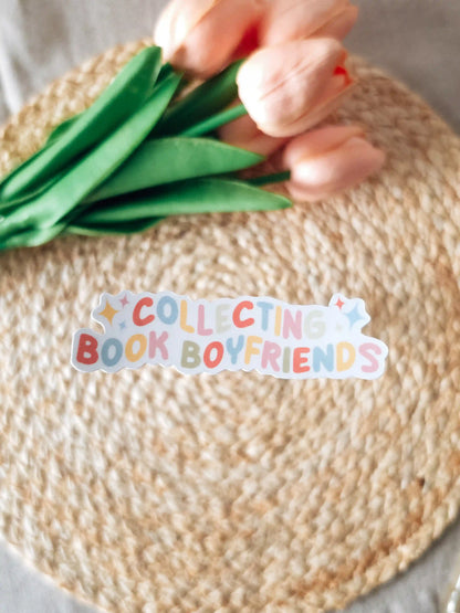 A die-cut sticker reading "Collecting Book Boyfriends" in multicolored rounded lettering with gold sparkle details, placed on a woven rattan mat with peach roses and green leaves resting in the upper corner.