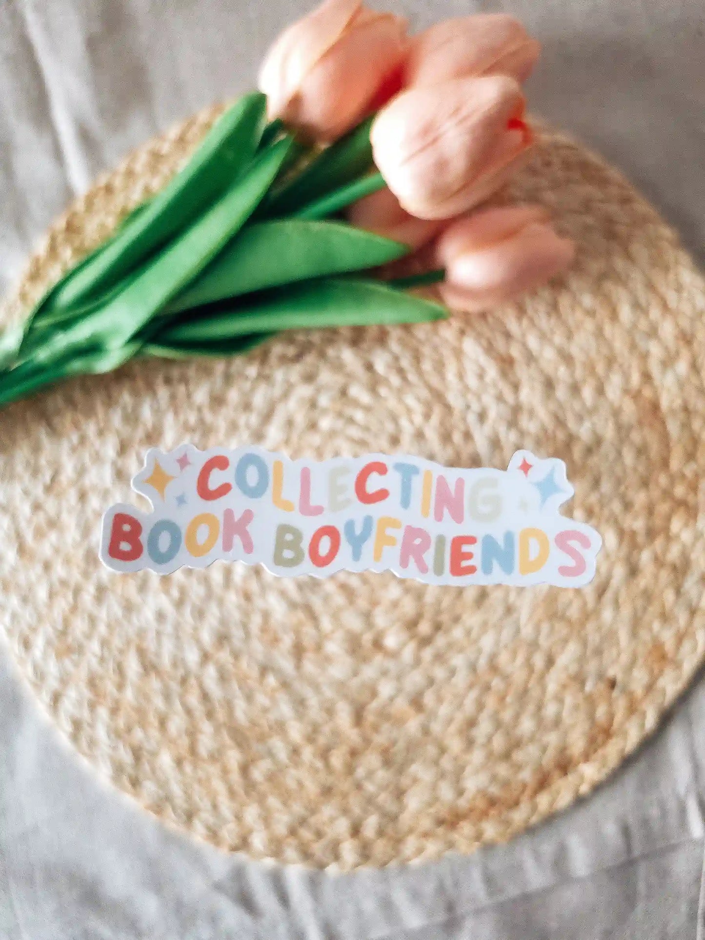 A die-cut sticker reading "Collecting Book Boyfriends" in multicolored rounded lettering with gold sparkle details, placed on a woven rattan mat with peach roses and green leaves resting in the upper corner.