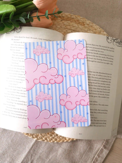 A E-Reader insert with a light blue and white striped background covered in large fluffy pink clouds and small blue star details, placed on an open book resting on a woven rattan mat. Peach roses and green rest in the upper corner.