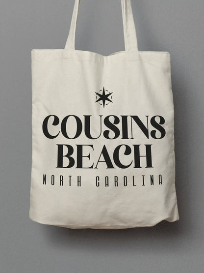 Cotton canvas tote bag with black "Cousins Beach, North Carolina" text, photographed on a solid gray background.