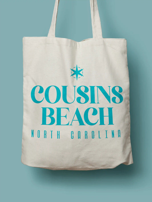 Cotton canvas tote bag with teal "Cousins Beach, North Carolina" text, photographed on a solid blue background.