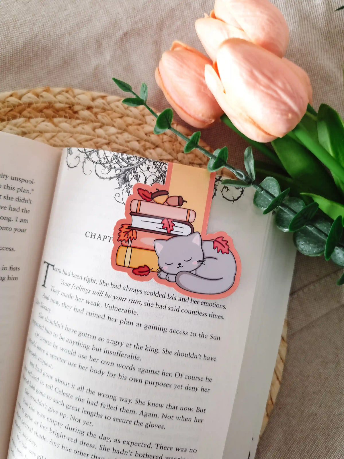 A die-cut magnetic bookmark featuring a sleeping grey cat with pink cheeks curled up beside a stack of illustrated books in pink, white, and yellow, decorated with red autumn leaves and small acorns, clipped onto the top corner of an open book resting on a woven rattan mat, with peach tulips and green eucalyptus sprigs in the upper right corner.