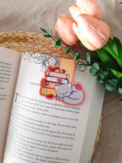 A die-cut magnetic bookmark featuring a sleeping grey cat with pink cheeks curled up beside a stack of illustrated books in pink, white, and yellow, decorated with red autumn leaves and small acorns, clipped onto the top corner of an open book resting on a woven rattan mat, with peach tulips and green eucalyptus sprigs in the upper right corner.