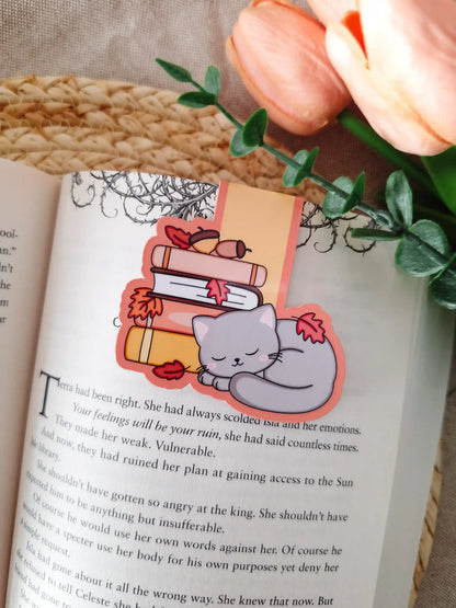A die-cut magnetic bookmark featuring a sleeping grey cat with pink cheeks curled up beside a stack of illustrated books in pink, white, and yellow, decorated with red autumn leaves and small acorns, clipped onto the top corner of an open book resting on a woven rattan mat, with peach tulips and green eucalyptus sprigs in the upper right corner.