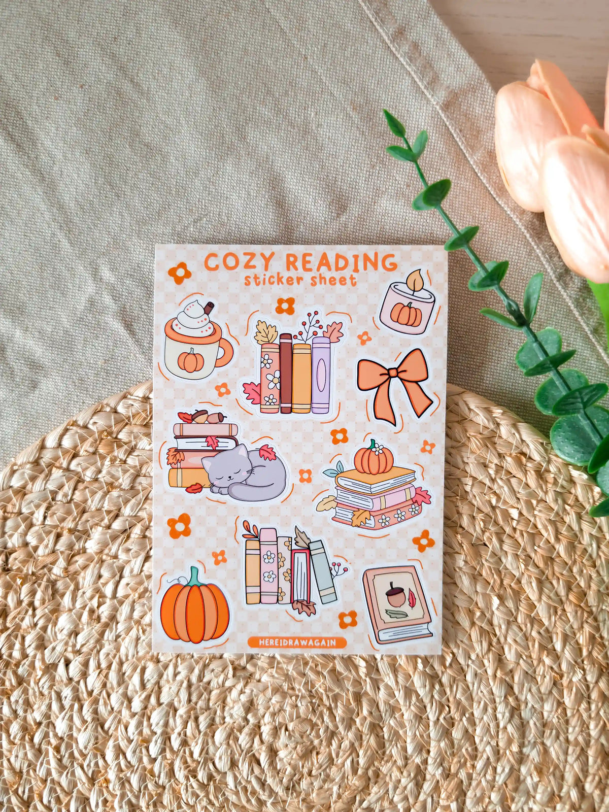 A glossy A6 sticker sheet titled "Cozy Reading Sticker Sheet" on a cream and orange checkered background, featuring nine illustrated autumn stickers including a pumpkin spice latte with whipped cream, two stacks of books with autumn leaves, a small pumpkin candle, an orange bow, a sleeping grey cat on books, a stack of pastel books with a pumpkin on top, an orange pumpkin, and a book with an acorn on the cover, displayed on a woven rattan mat with green eucalyptus sprigs and a peach tulip in the upper right