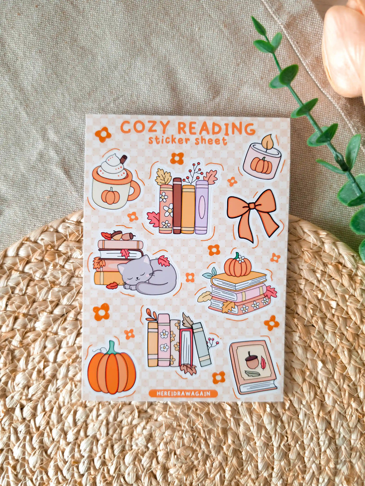 A glossy A6 sticker sheet titled "Cozy Reading Sticker Sheet" on a cream and orange checkered background, featuring nine illustrated autumn stickers including a pumpkin spice latte with whipped cream, two stacks of books with autumn leaves, a small pumpkin candle, an orange bow, a sleeping grey cat on books, a stack of pastel books with a pumpkin on top, an orange pumpkin, and a book with an acorn on the cover, displayed on a woven rattan mat with green eucalyptus sprigs and a peach tulip in the upper right