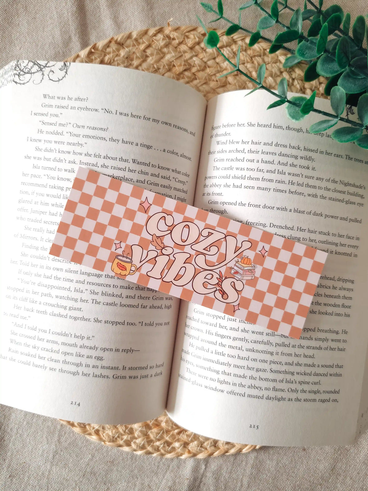 A horizontal laminated bookmark with a terracotta and white checkered pattern and the text "cozy vibes" in retro-style lettering, decorated with small illustrations of a steaming mug, autumn leaves, and a stack of books with a pumpkin, resting on an open book on a woven rattan mat, with green eucalyptus sprigs in the upper right corner.