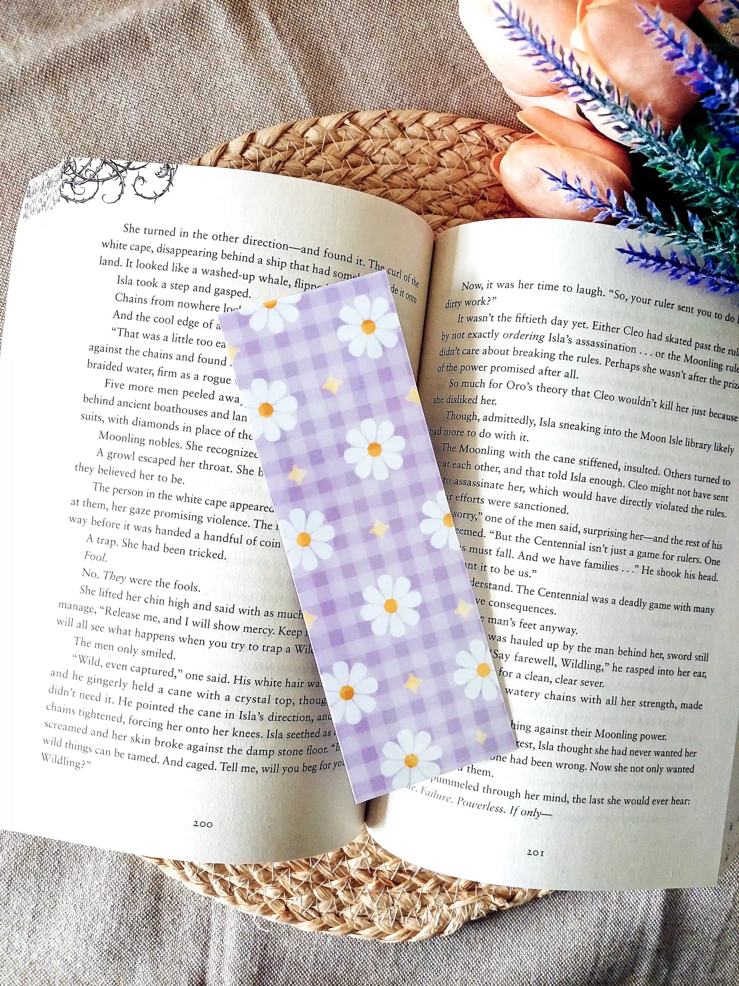 Bookmark featuring a purple gingham pattern with white daisies and small yellow star details, displayed on an open book over a woven mat. 