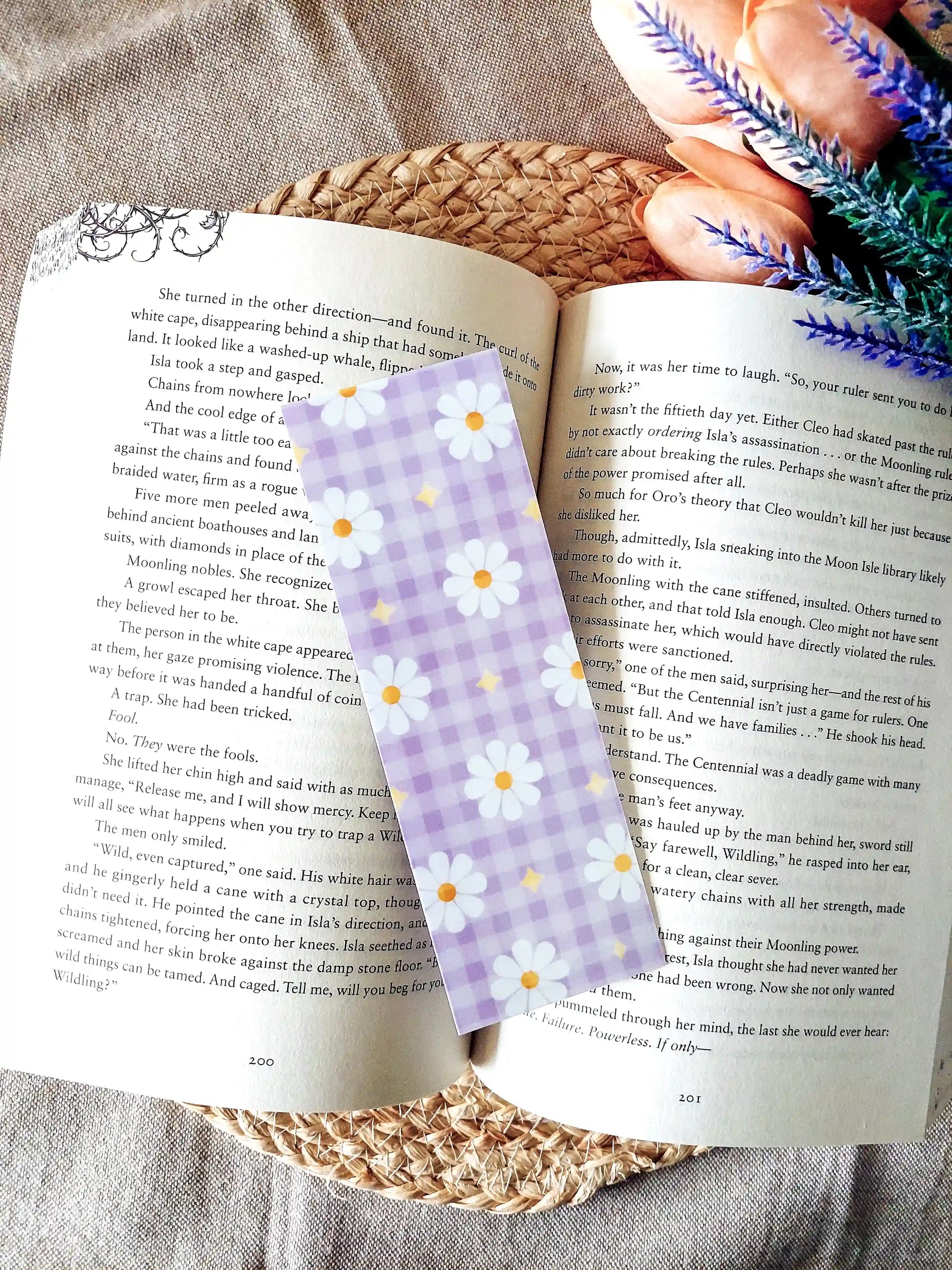 Bookmark featuring a purple gingham pattern with white daisies and small yellow star details, displayed on an open book over a woven mat. 