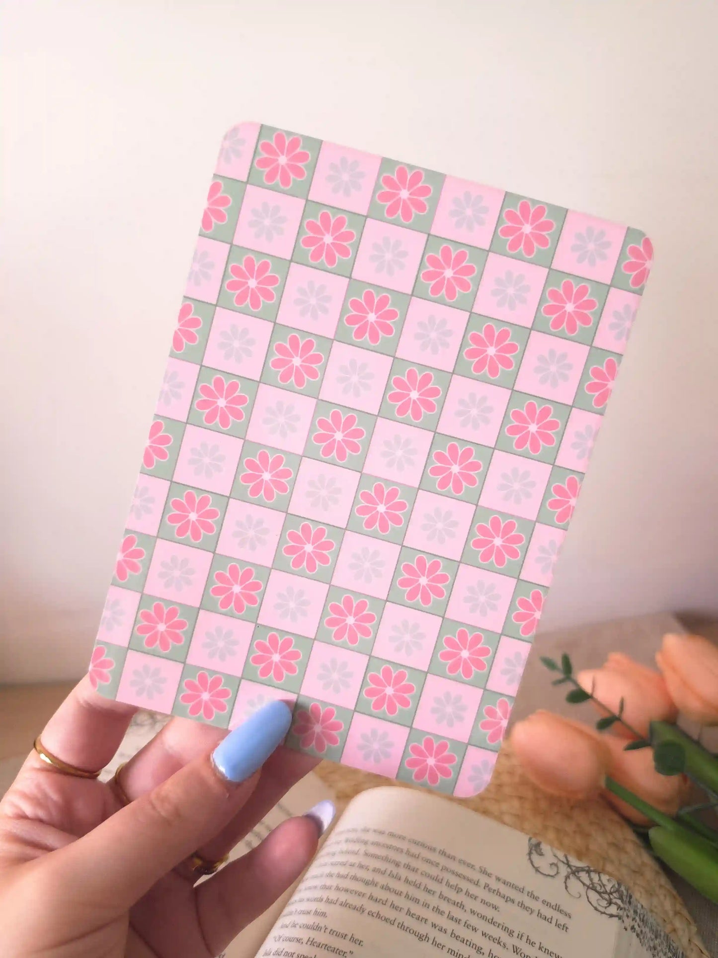 A hand holding an e-reader insert with a pink and sage green checkerboard pattern, each square featuring a pink or blue daisy illustration.