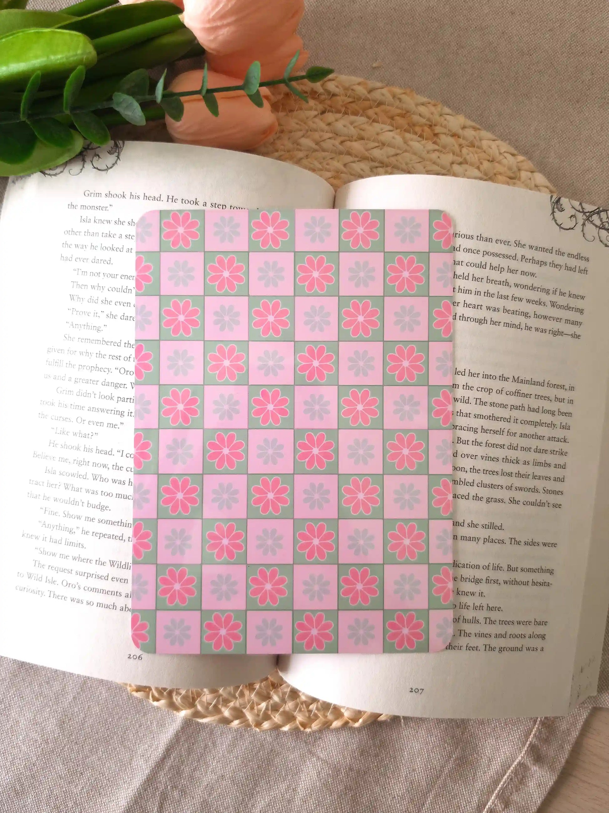 An e-reader insert with a pink and sage green checkerboard pattern, each square featuring a pink or blue daisy illustration, placed on an open book resting on a woven rattan mat. 