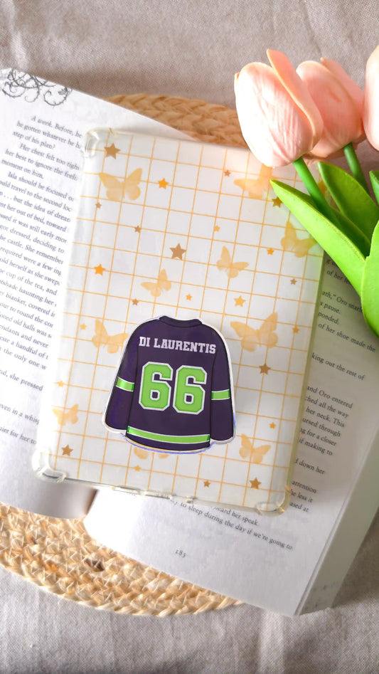 Dark navy hockey jersey magnetic grip with green “Di Laurentis 66" attached to an e-reader with a cream grid insert of golden butterflies and stars, lying on an open book on a woven rattan mat, with peach tulips on the right.