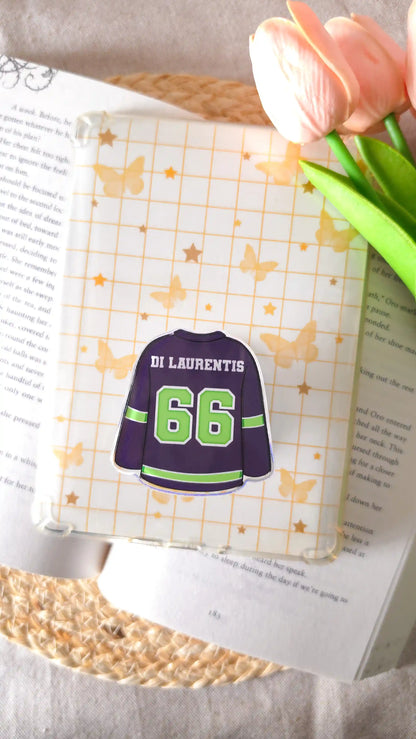 Dark navy hockey jersey magnetic grip with green “Di Laurentis 66" attached to an e-reader with a cream grid insert of golden butterflies and stars, lying on an open book on a woven rattan mat, with peach tulips on the right.