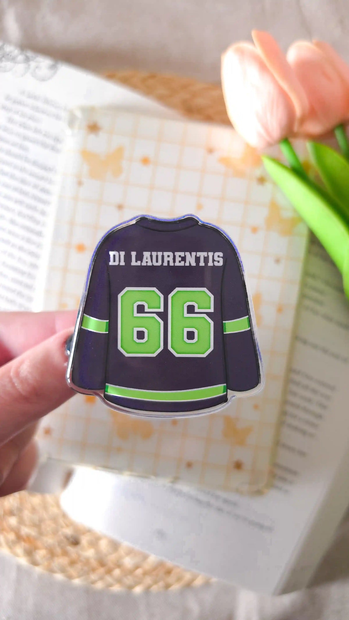 Hand holding a dark navy hockey jersey magnetic grip with “Di Laurentis 66”, in front of a cream magnetic bookmark with a butterfly and grid pattern, an open book, pink tulips and a woven rattan mat.