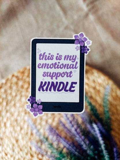 Vinyl sticker of a black Kindle with coral and pink flowers, featuring the phrase "This is my emotional support Kindle", photographed on a neutral woven background.