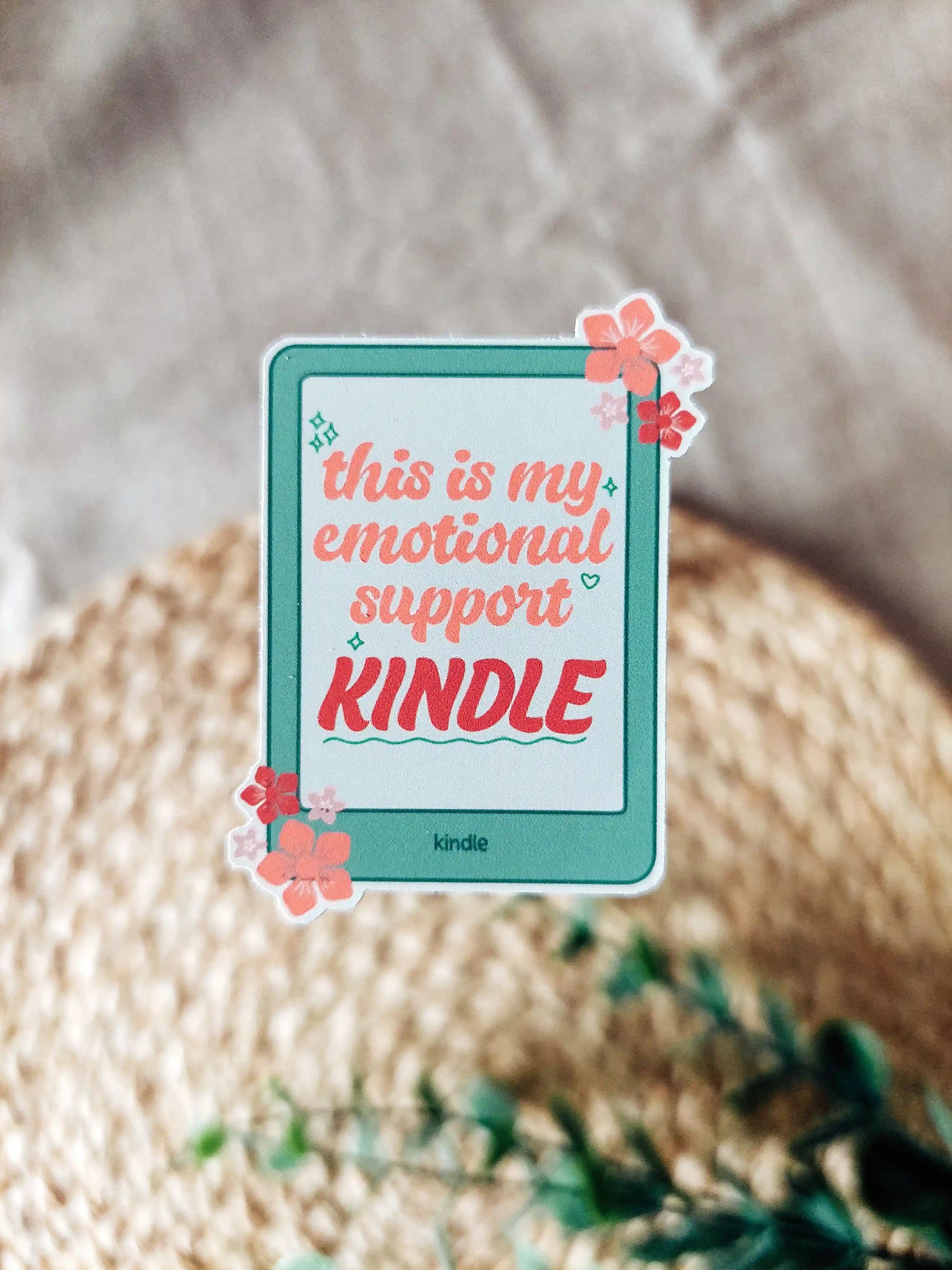 Vinyl sticker of a matcha green Kindle with coral and pink flowers, featuring the phrase "This is my emotional support Kindle", photographed on a neutral woven background.
