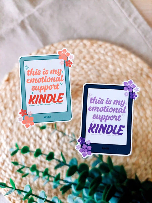 Two stickers of a Kindle with the phrase This is my emotional support Kindle" in green and black color variants, photographed on a woven mat.