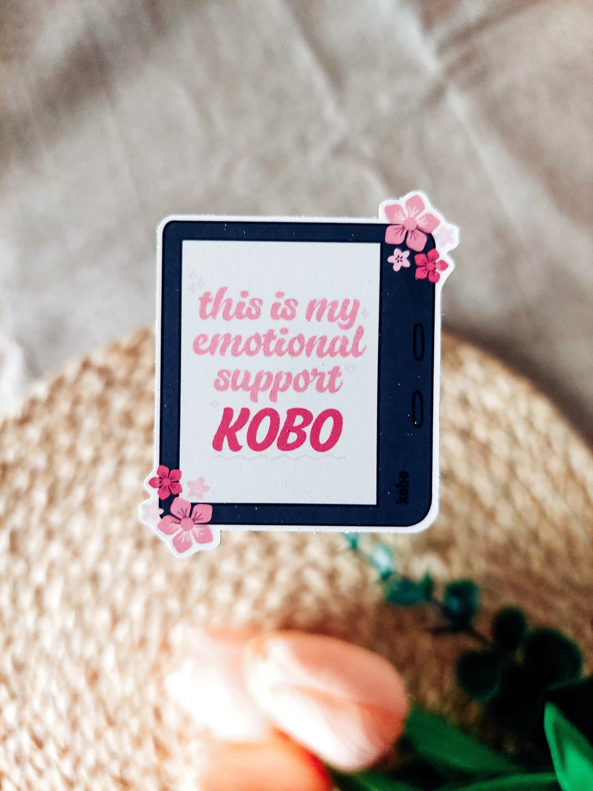 Vinyl sticker of a black Kobo with pink floral details, featuring the phrase "This is my emotional support Kobo", photographed on a neutral woven background.