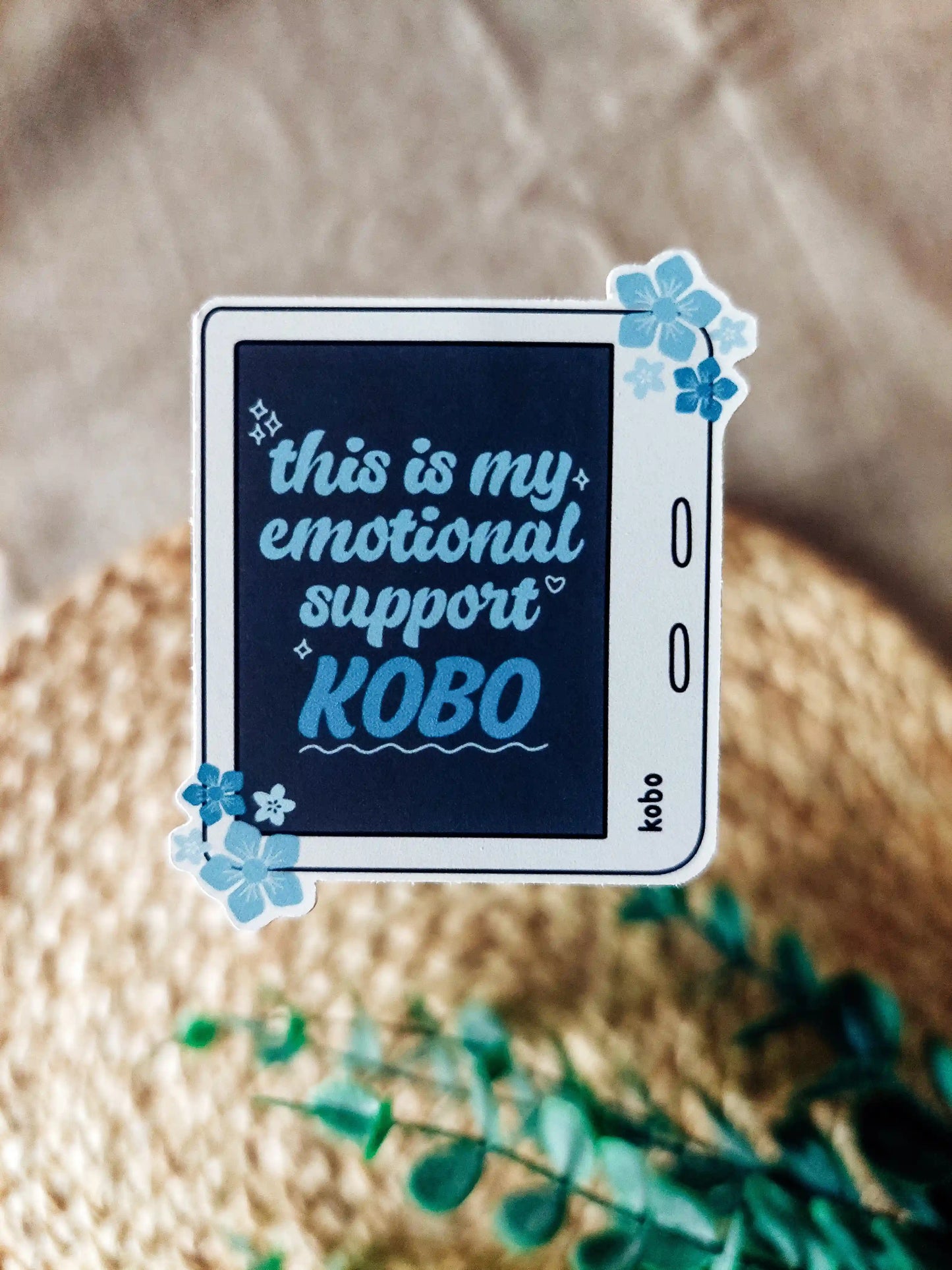 Vinyl sticker of a white Kobo with blue floral details, featuring the phrase "This is my emotional support Kobo", photographed on a neutral woven background.