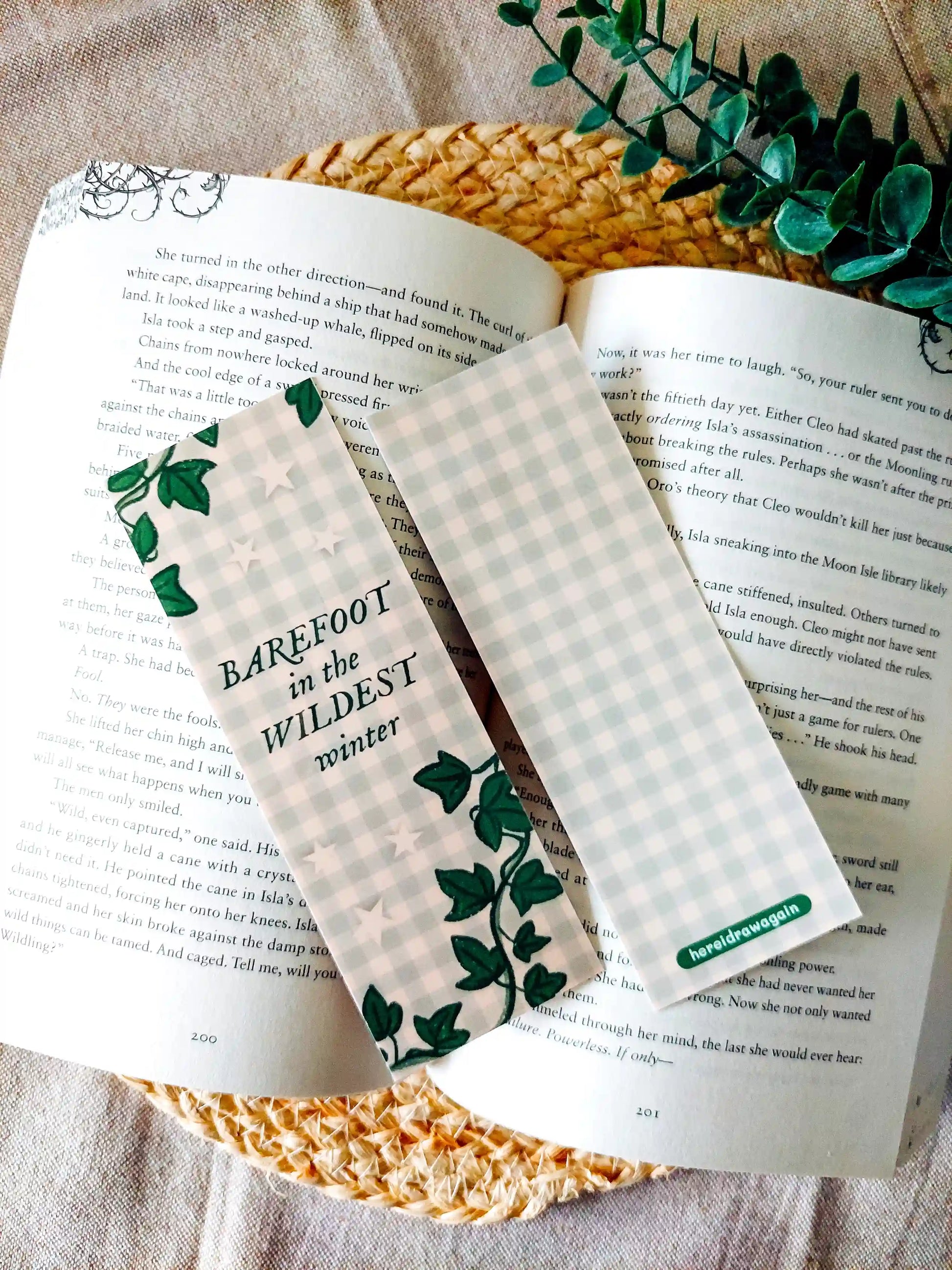 Bookmark inspired by Taylor Swift's Evermore featuring ivy illustrations and green gingham background, with the lyric "barefoot in the wildest winter", displayed on an open book over a woven mat surface decorative plants.