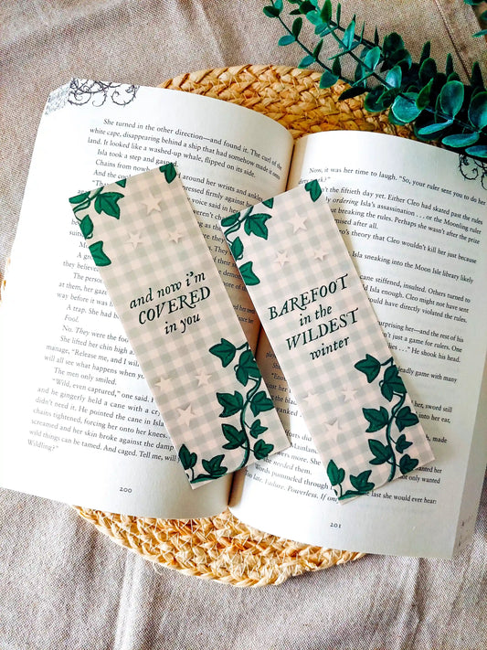 Bookmarks inspired by Taylor Swift's Evermore featuring ivy illustrations and green gingham backgrounds, with the lyrics "and now I'm covered in you" and "barefoot in the wildest winter", displayed on an open book over a woven mat surface decorative plants.
