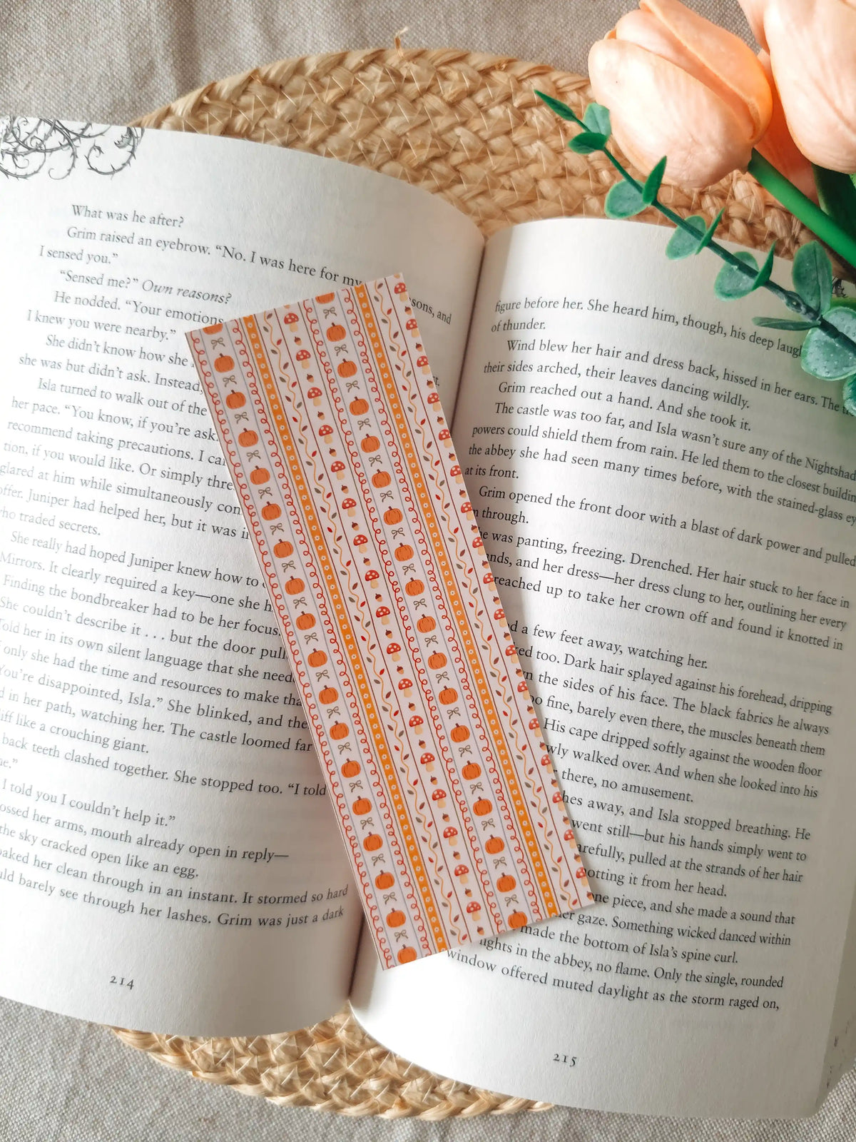 A laminated bookmark with a cream background and vertical orange stripes, featuring illustrated pumpkins, red mushrooms with white dots, autumn maple leaves, and small bows in warm orange tones, resting on an open book on a woven rattan mat, with peach tulips and green eucalyptus sprigs in the upper right corner.