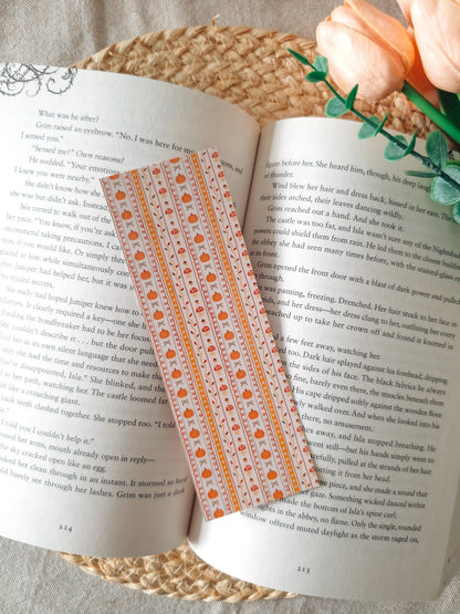 A laminated bookmark with a cream background and vertical orange stripes, featuring illustrated pumpkins, red mushrooms with white dots, autumn maple leaves, and small bows in warm orange tones, resting on an open book on a woven rattan mat, with peach tulips and green eucalyptus sprigs in the upper right corner.