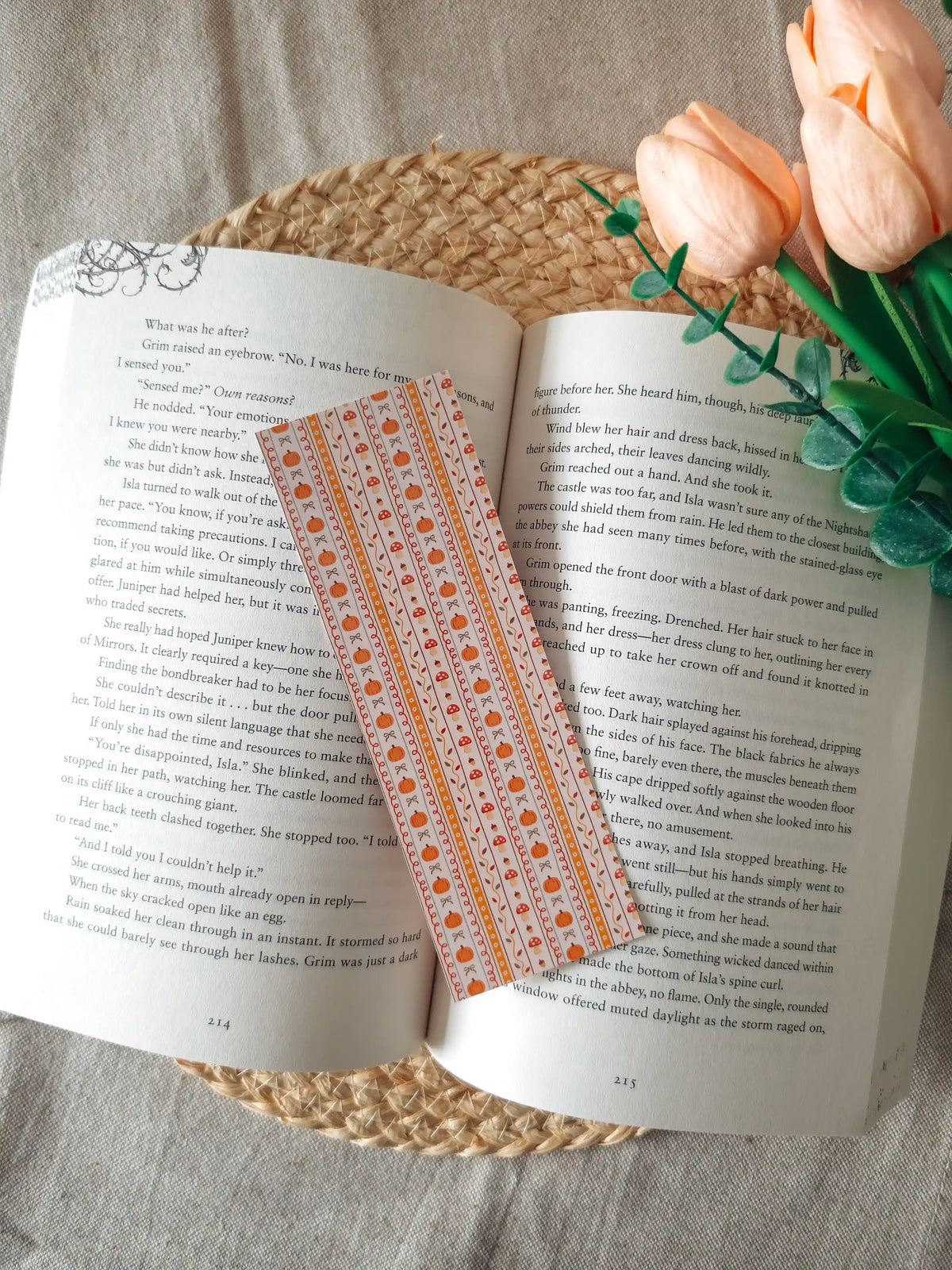 A laminated bookmark with a cream background and vertical orange stripes, featuring illustrated pumpkins, red mushrooms with white dots, autumn maple leaves, and small bows in warm orange tones, resting on an open book on a woven rattan mat, with peach tulips and green eucalyptus sprigs in the upper right corner.