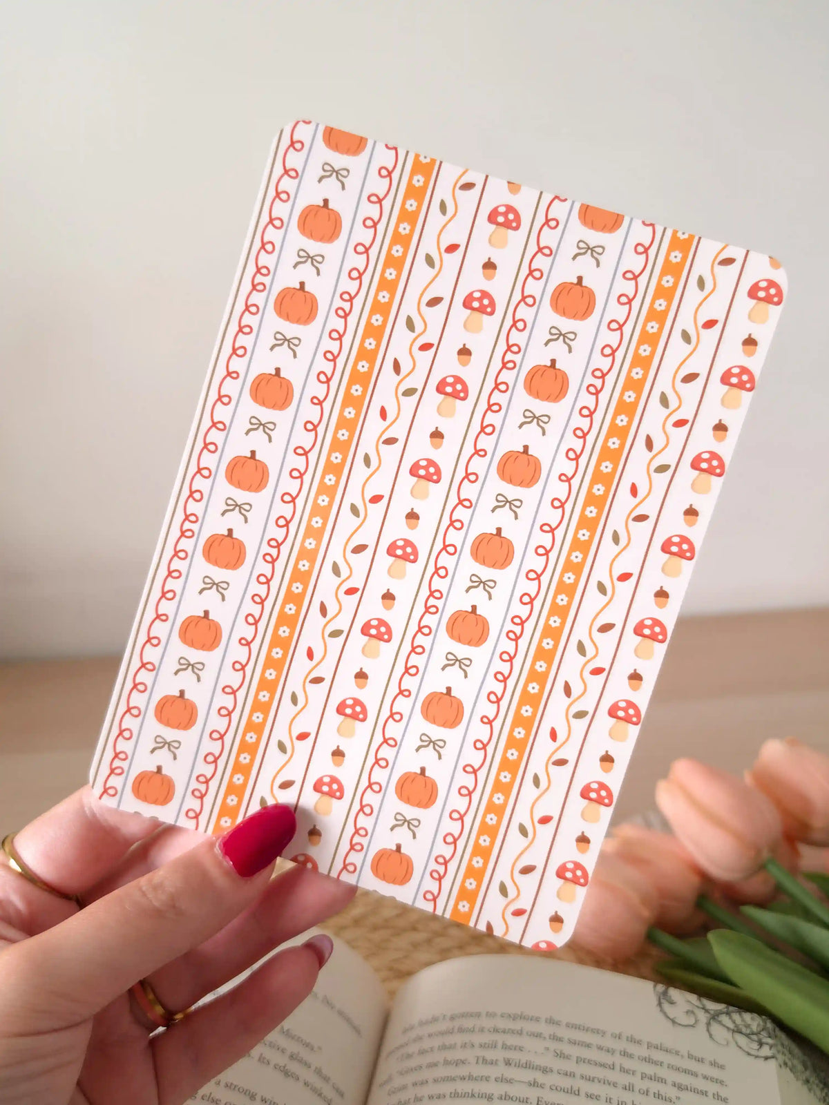 A laminated e-reader insert with a cream and orange vertical stripe pattern featuring illustrated pumpkins, red mushrooms with white dots, acorns, autumn leaves, and small bows in warm orange tones, resting on an open book on a woven rattan mat, with green eucalyptus sprigs in the upper left corner.