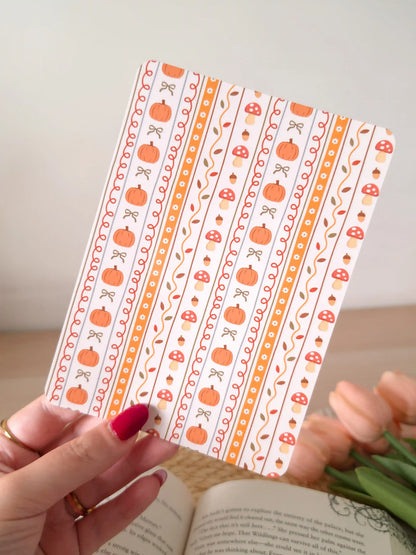 A laminated e-reader insert with a cream and orange vertical stripe pattern featuring illustrated pumpkins, red mushrooms with white dots, acorns, autumn leaves, and small bows in warm orange tones, resting on an open book on a woven rattan mat, with green eucalyptus sprigs in the upper left corner.