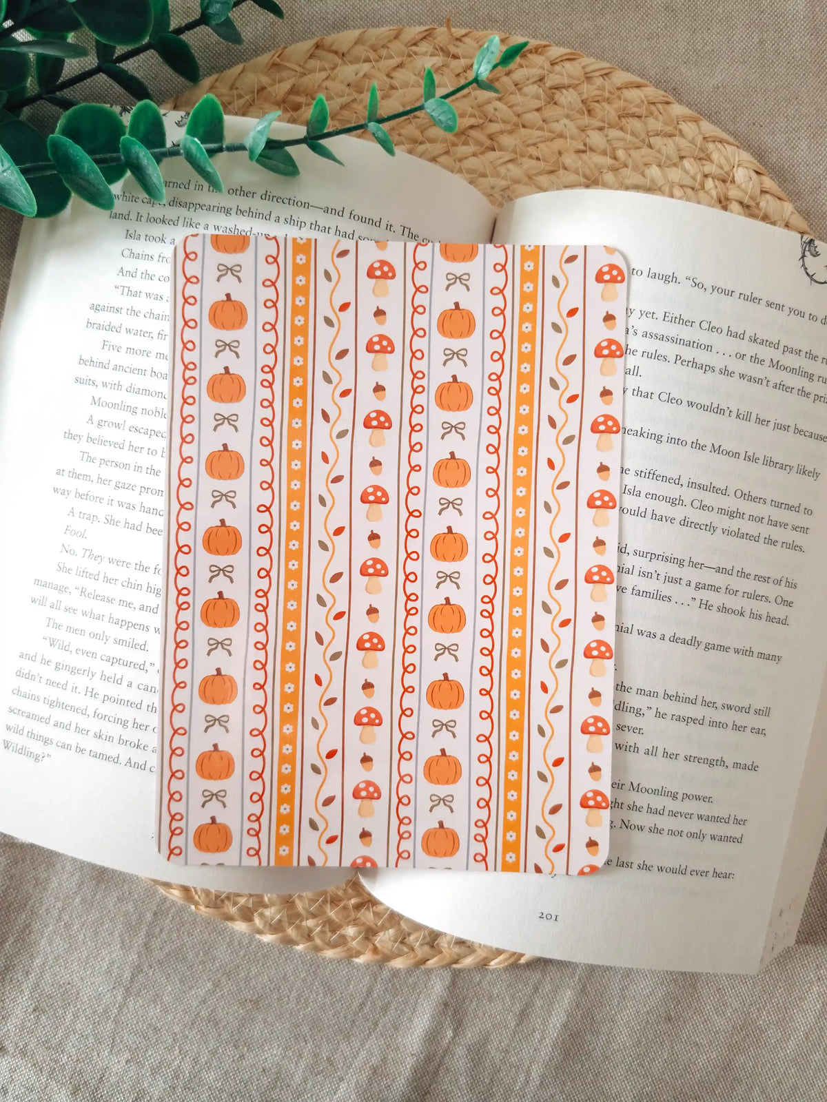 A laminated e-reader insert with a cream and orange vertical stripe pattern featuring illustrated pumpkins, red mushrooms with white dots, acorns, autumn leaves, and small bows in warm orange tones, resting on an open book on a woven rattan mat, with green eucalyptus sprigs in the upper left corner.