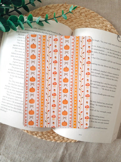 A laminated e-reader insert with a cream and orange vertical stripe pattern featuring illustrated pumpkins, red mushrooms with white dots, acorns, autumn leaves, and small bows in warm orange tones, resting on an open book on a woven rattan mat, with green eucalyptus sprigs in the upper left corner.