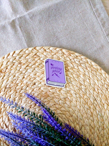 Vinyl sticker featuring a purple book with bats and a sword illustration on the cover and the text "Fantasy Girlie", photographed on a neutral fabric background with woven mat.