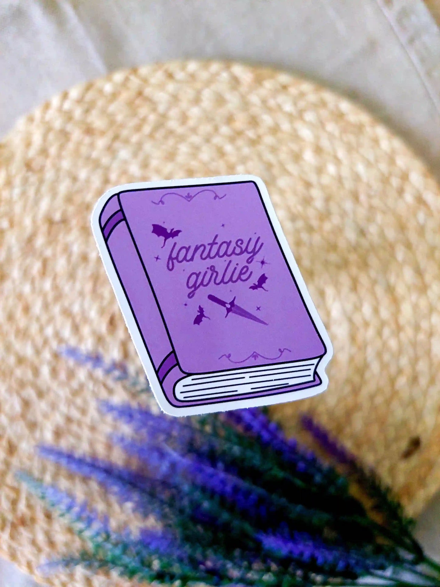 Vinyl sticker featuring a purple book with bats and a sword illustration on the cover and the text "Fantasy Girlie", photographed on a neutral fabric background with woven mat.