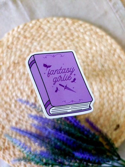 Vinyl sticker featuring a purple book with bats and a sword illustration on the cover and the text "Fantasy Girlie", photographed on a neutral fabric background with woven mat.