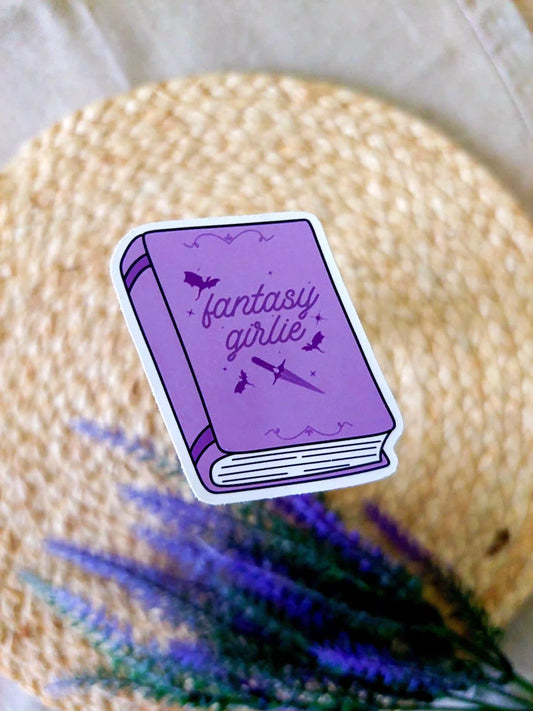 Vinyl sticker featuring a purple book with bats and a sword illustration on the cover and the text "Fantasy Girlie", photographed on a neutral fabric background with woven mat.
